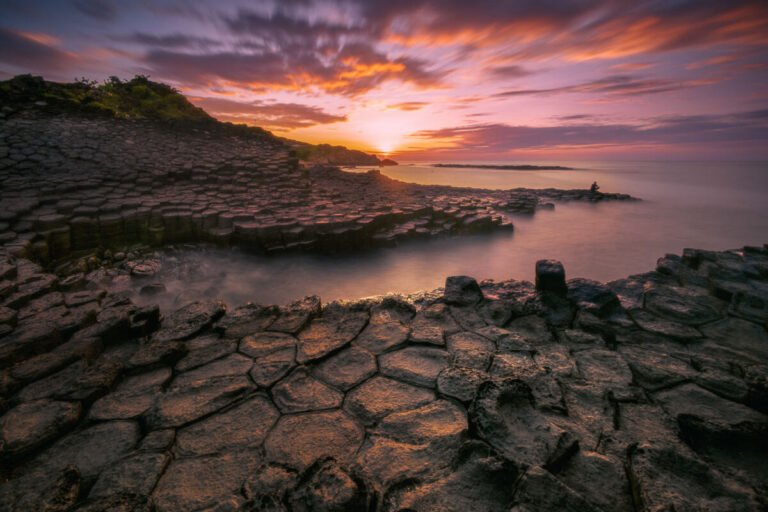 Dark hexagonal basalt rocks lead toward a calm sea under a glowing orange and purple sunset sky.