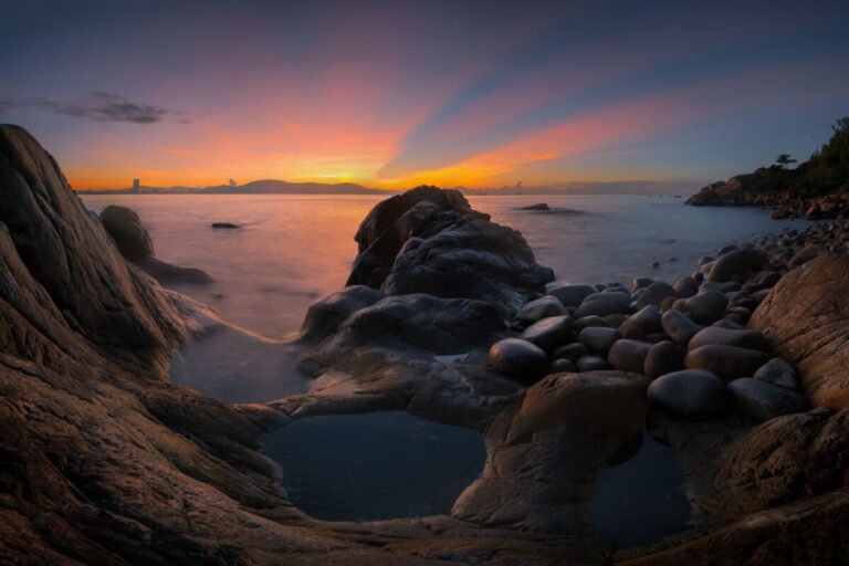A rocky cove with rounded boulders around a still pool faces the sea as the sun sets beyond distant hills.