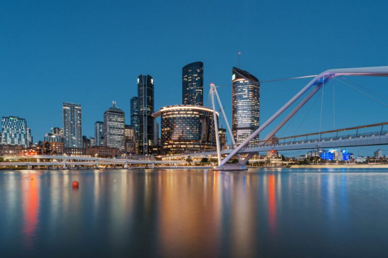 A pedestrian footbridge crosses the river toward Brisbane's lit skyline, with colourful reflections on the water.