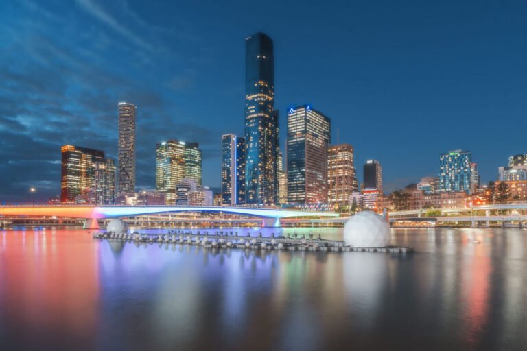 Brisbane's illuminated towers and a lit bridge reflect in smooth, colourful bands on the river at dusk.