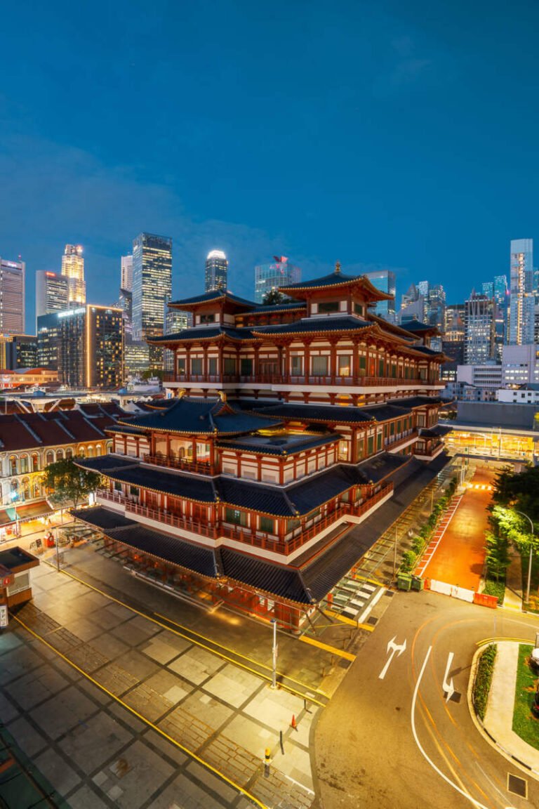 An ornate red multi-tiered temple in Singapore's Chinatown glows at blue hour, with city towers rising behind it.