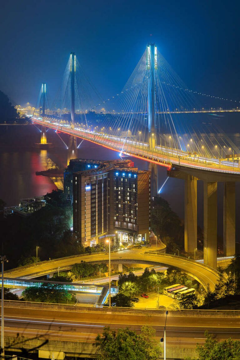 An illuminated cable-stayed bridge spans the water at blue hour above a glowing hotel and curving highway.