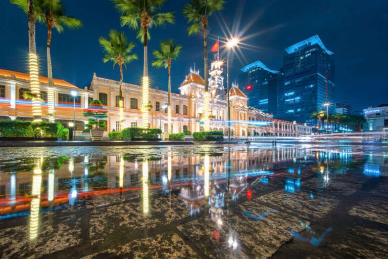 An ornate lit colonial building with palm trees and modern towers reflects in a wet plaza at night.