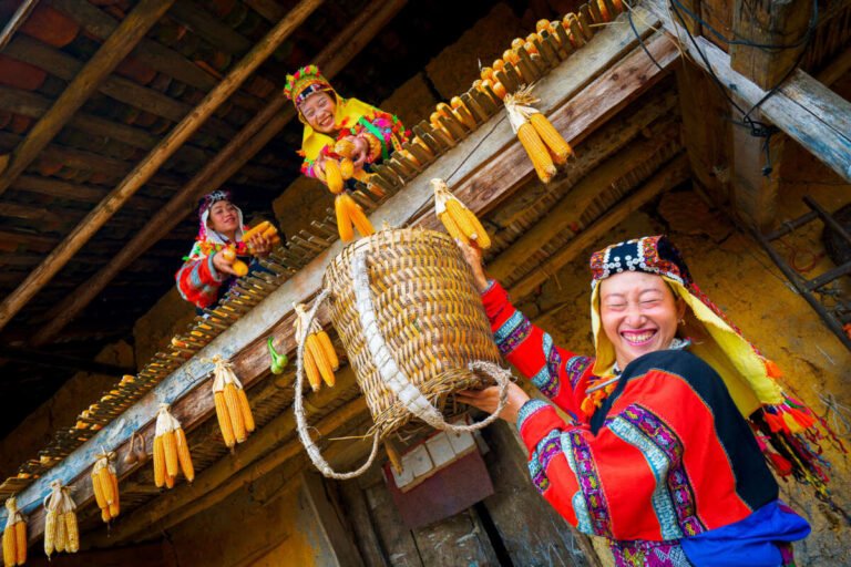 Women in bright traditional dress hang ears of golden corn and a woven basket from the wooden eaves of a house.