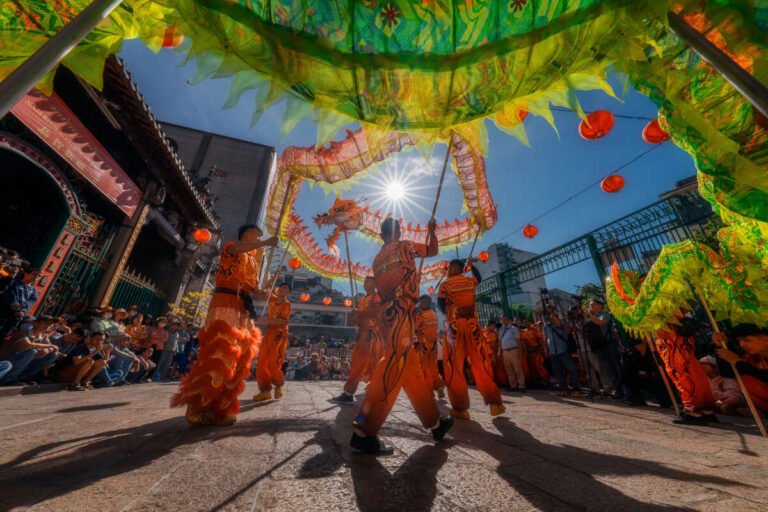 Performers in orange costumes dance beneath a green dragon and red lanterns, backlit by a bright sun.