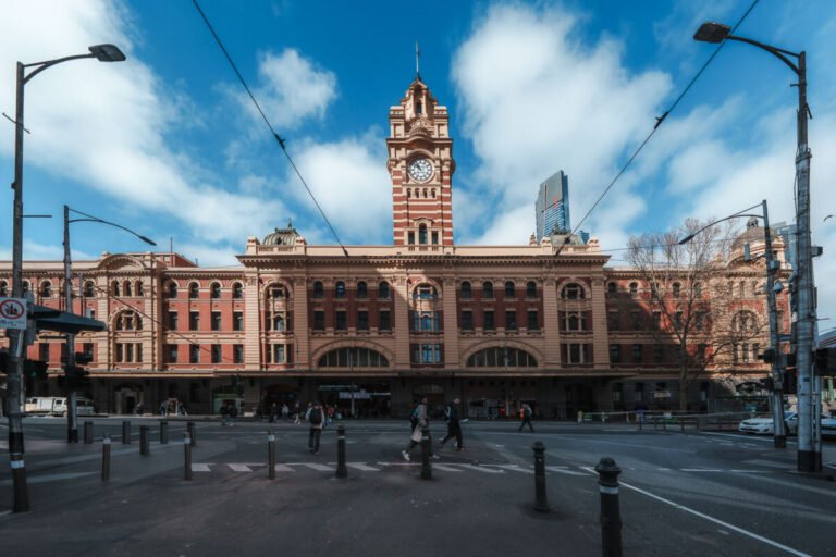 The ornate facade and clock tower of Flinders Street Station in Melbourne under a bright blue sky with white clouds.