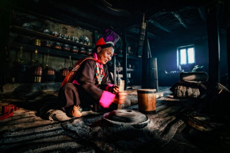A woman in a traditional headdress kneels at a stone hand mill in a dim kitchen lit by a small window.