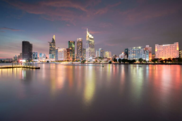 The lit skyline of Ho Chi Minh City at dusk reflects in colourful bands across a calm river.