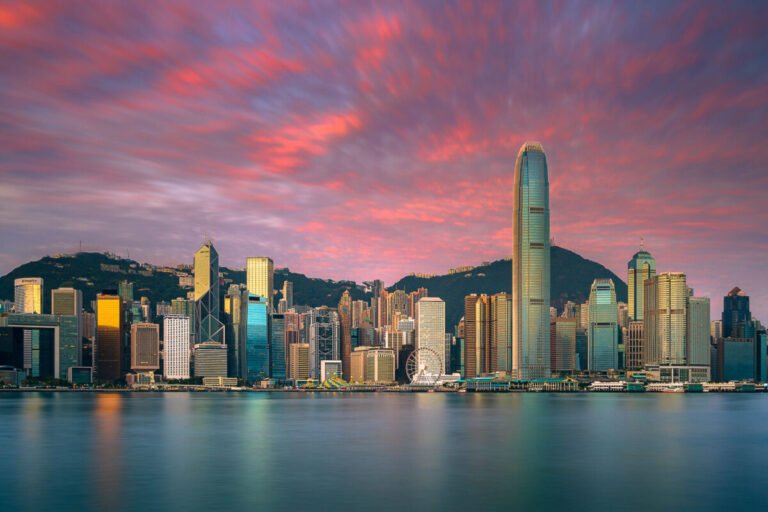 The Hong Kong Island skyline lines Victoria Harbour beneath vivid pink and orange clouds at sunset.