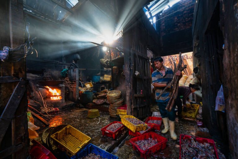 Beams of light stream through a dark wooden workshop where a man works beside a fire and red crates.