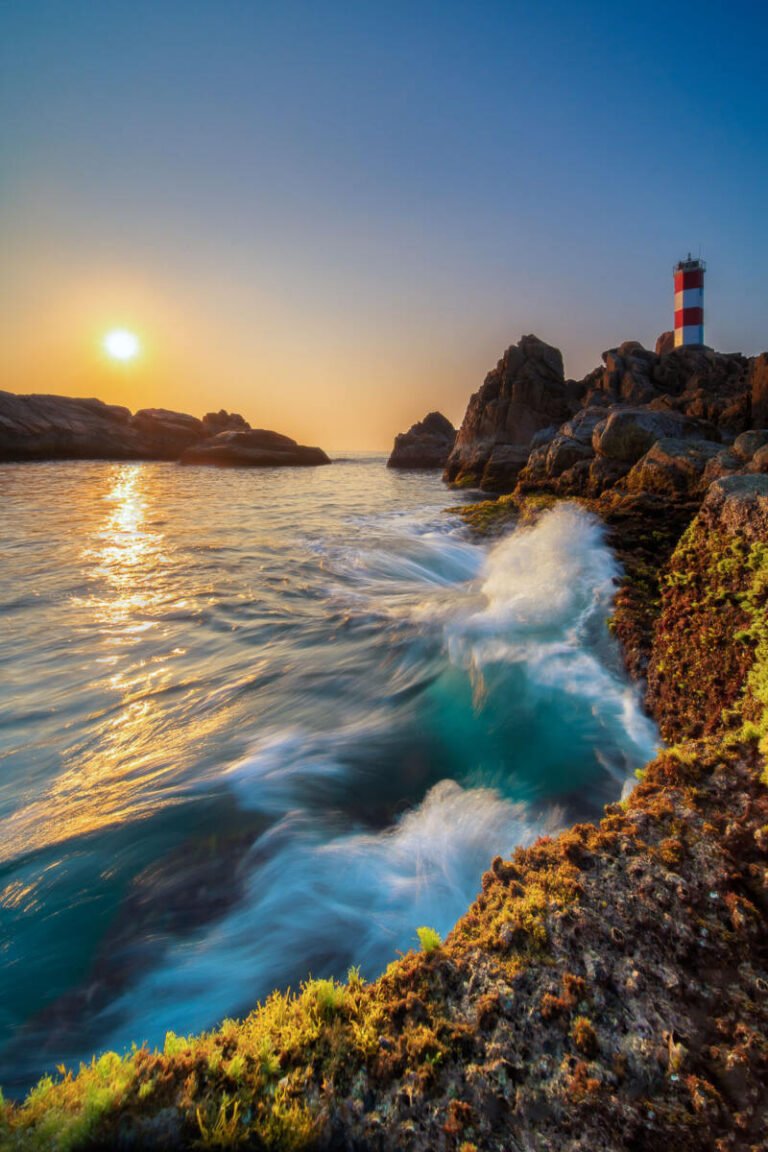 Waves crash against a rocky headland topped by a red and white lighthouse as the sun rises over the sea.