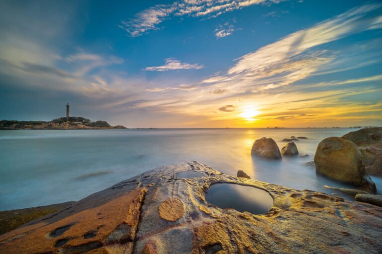 A rocky shoreline with weathered boulders and a tidal pool faces a rising sun, with a distant lighthouse on the headland.