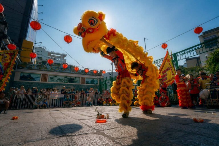 A golden lion dancer rears up in a lantern-hung courtyard as a crowd watches under a bright blue sky.