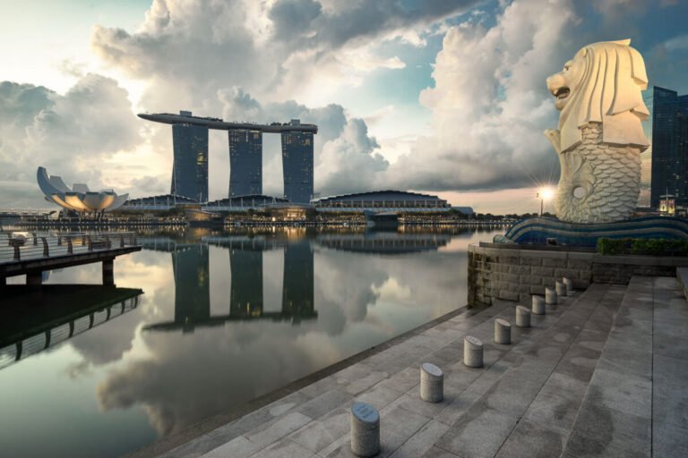 The Merlion statue stands by still water reflecting Marina Bay Sands beneath towering white clouds.