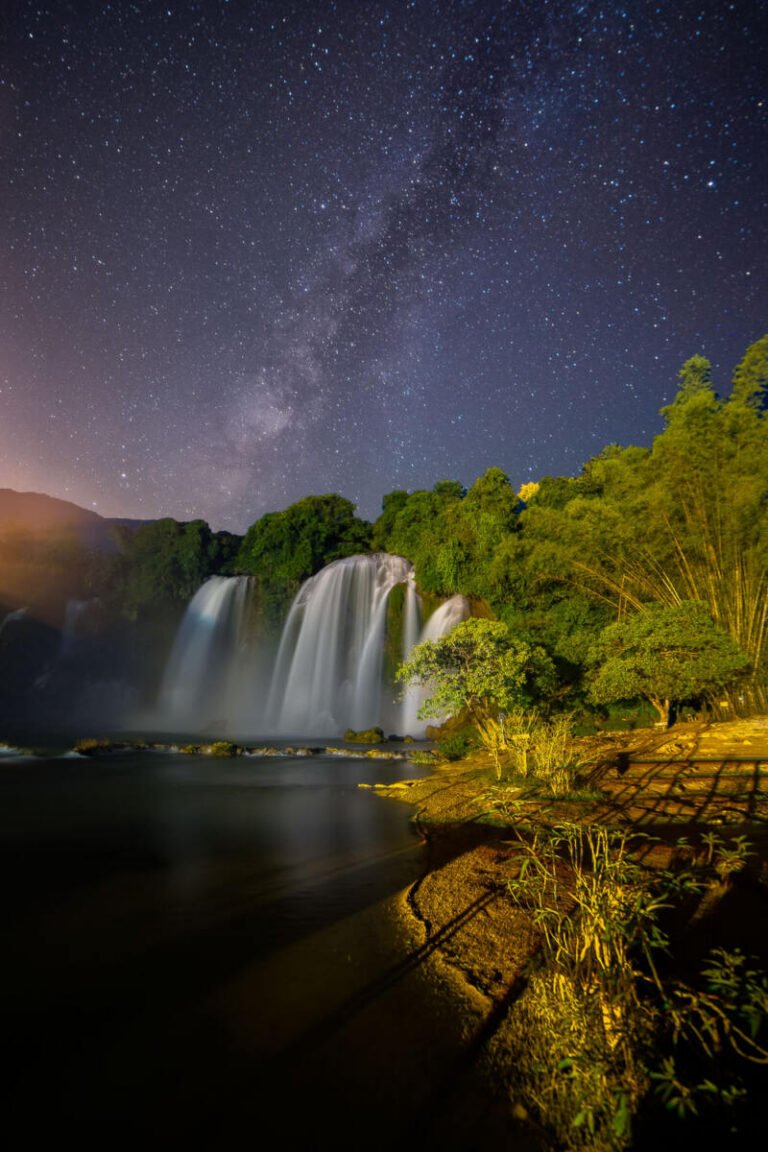 A wide waterfall pours into a dark pool beneath a starry night sky with the Milky Way rising above lush trees.