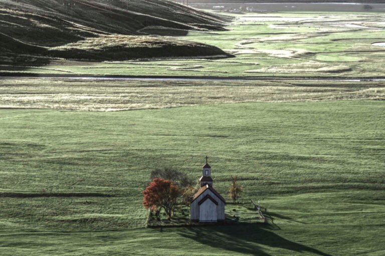 Lone chapel and red tree on green pasture with a bright braided river plain behind