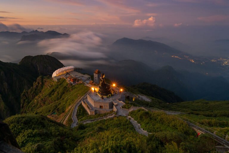 A shrine and winding stairway crown a green mountain summit above a sea of evening cloud and distant lights.