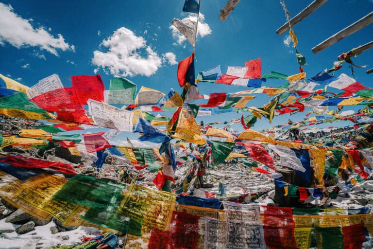 Strings of colourful prayer flags flutter across a mountain pass with patches of snow under a blue sky.