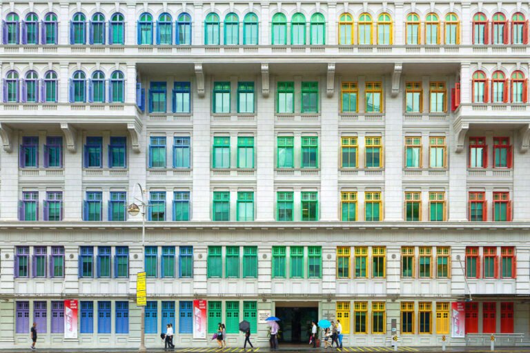 A long white colonial-style building facade with rows of windows painted in bright rainbow colours above a pedestrian street.