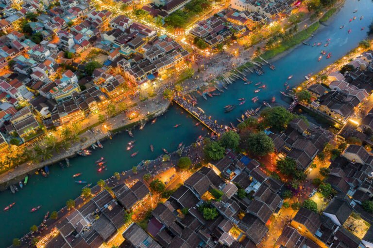 Aerial evening view of a river lined with small boats, crossed by a bridge and flanked by densely packed lit houses.