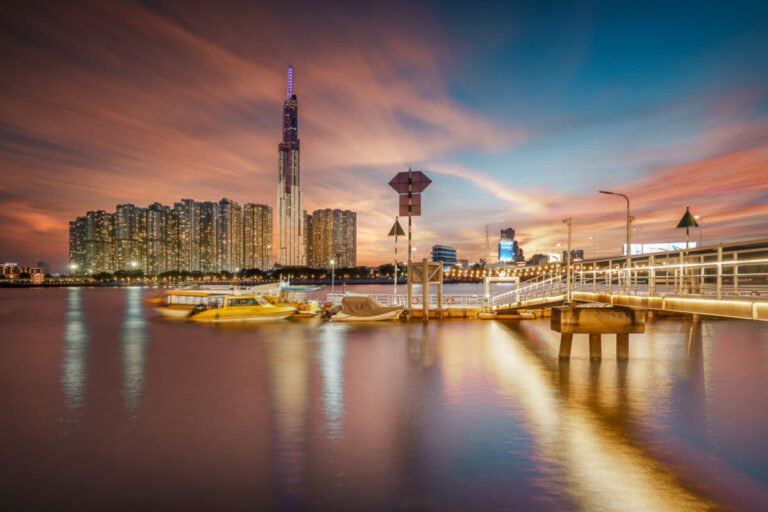 Sunset view across a calm river toward a tall skyscraper and apartment towers, with a pier and glowing reflections.