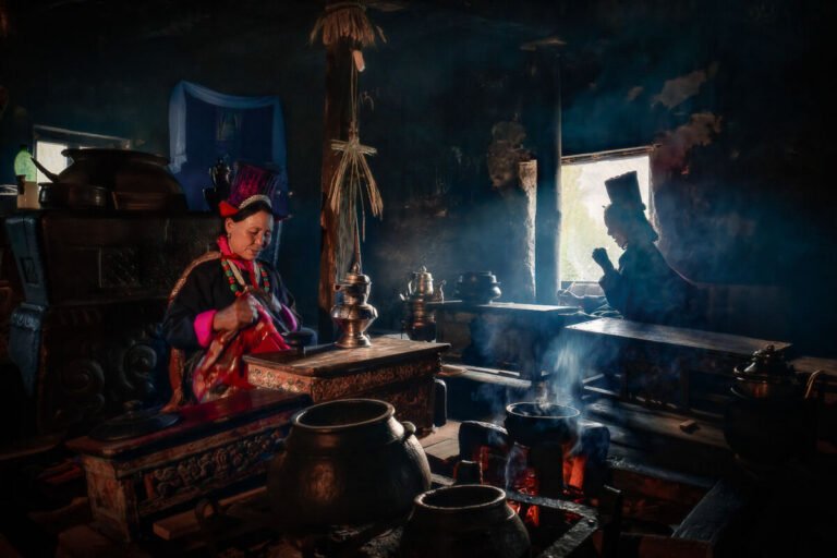 A woman in traditional dress tends a stove in a dark, smoky kitchen lined with copper pots and window light.