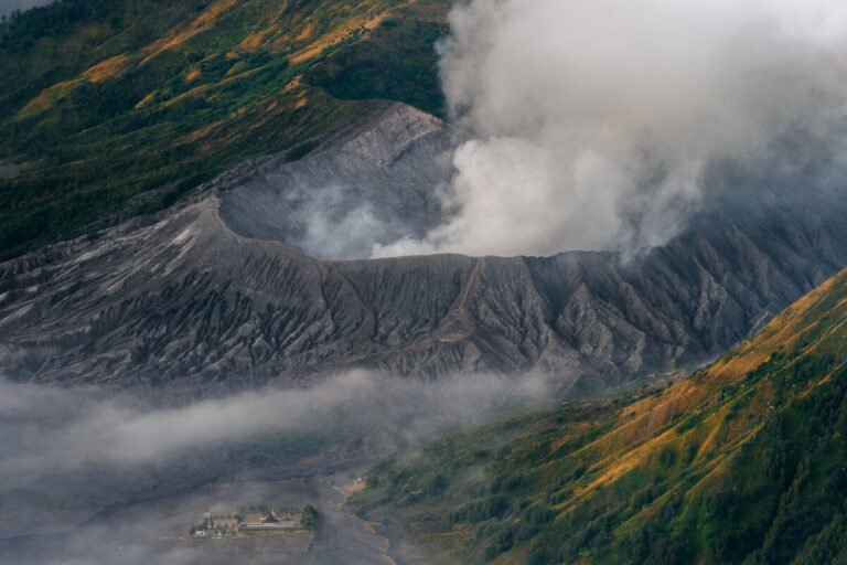 Steam billows from a vast grey volcanic crater surrounded by ridged slopes and drifting cloud.