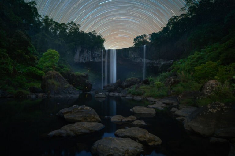 Circular star trails arc above a waterfall pouring into a rocky pool surrounded by dark forested cliffs.