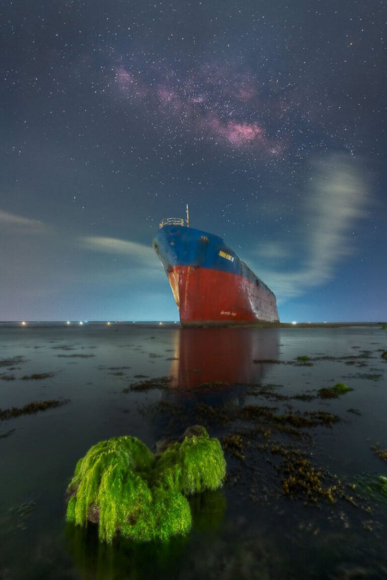 Vertical night view of a large stranded ship in calm water beneath the Milky Way, with green moss-covered rocks in front.