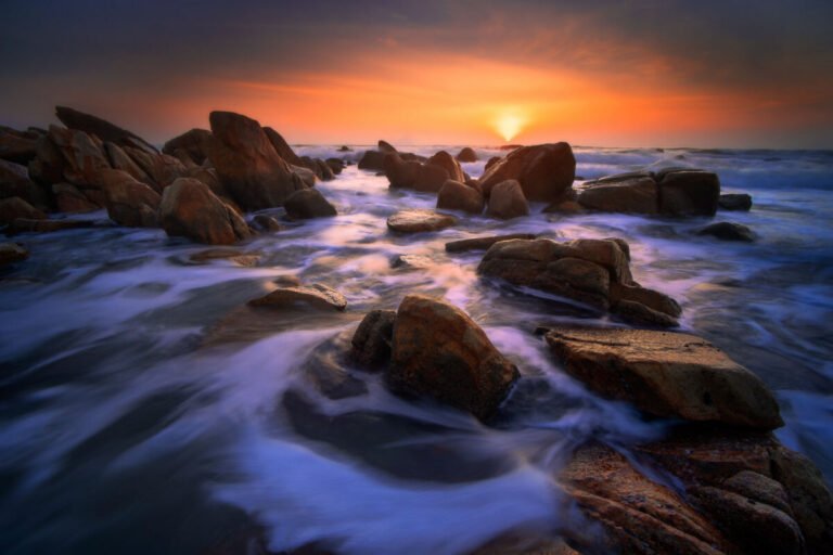 Long-exposure view of waves swirling around dark coastal boulders as the sun sets on the horizon.