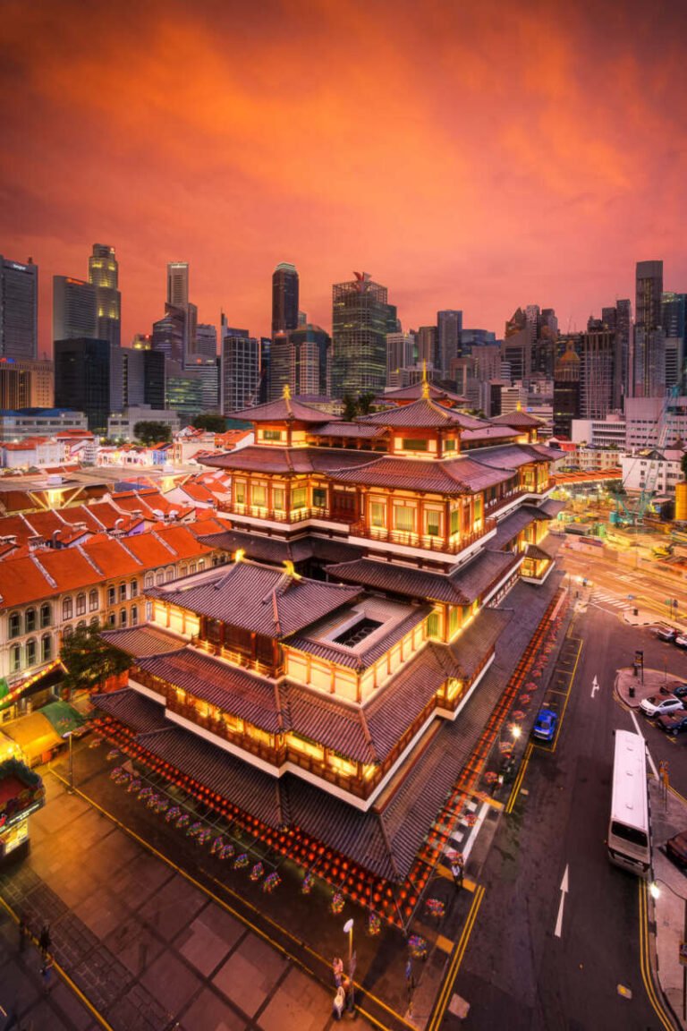 An ornate multi-tiered red temple lit at dusk, surrounded by streets and modern skyscrapers under a fiery orange sky.
