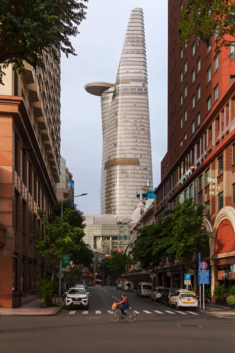 Vertical street view toward a tall tapered skyscraper rising between buildings, with traffic and a cyclist below.