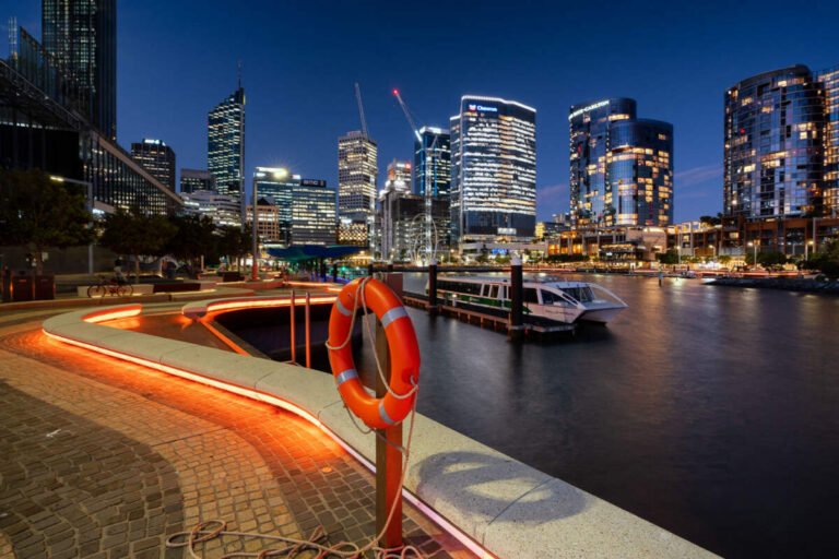 An orange-lit curving promenade with a life ring overlooks a harbour and illuminated city towers at blue hour.