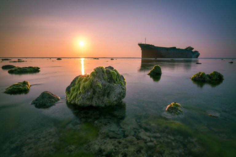 Mossy rocks rise from calm shallow sea at sunrise, with a rusting shipwreck on the horizon to the right.